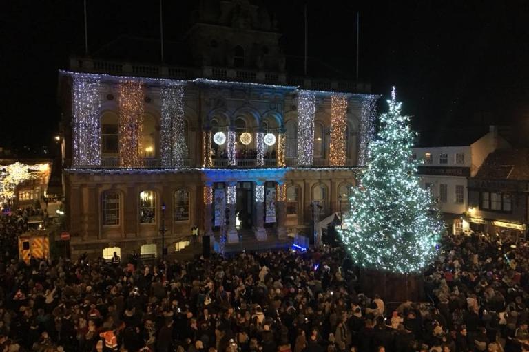 A town square with crowds, and a Christmas tree and Town Hall decorated in Christmas lights