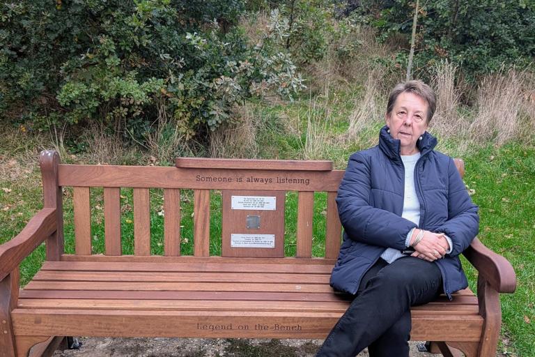 Woman in a blue jacket and dark trousers sitting on a wooden park bench 