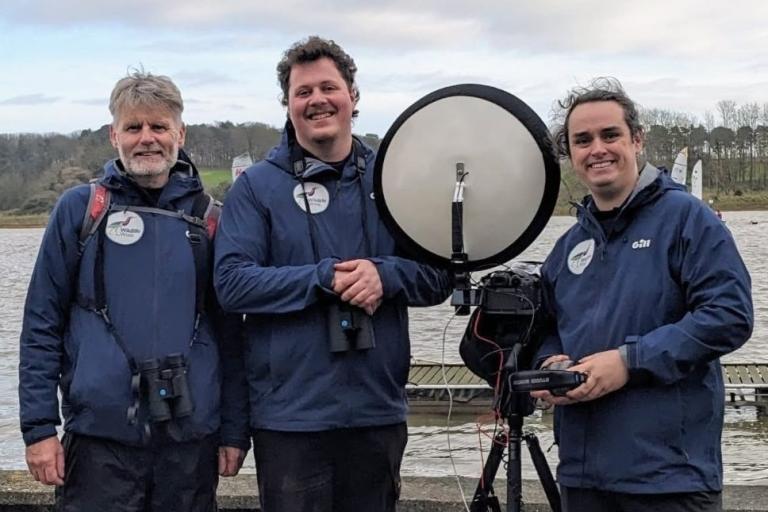 Three men with monitoring equipment standing beside a river estuary