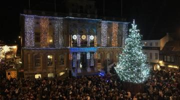 A town square with crowds, and a Christmas tree and Town Hall decorated in Christmas lights