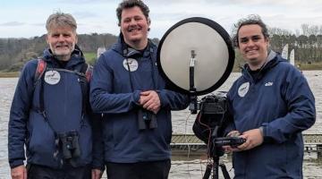 Three men with monitoring equipment standing beside a river estuary