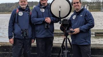 Three men with monitoring equipment standing beside a river estuary